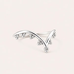 May include: A silver-colored ring with a V-shaped design. The ring features small, round bead-like embellishments along the curved lines of the V. The ring is set against a soft, neutral background.