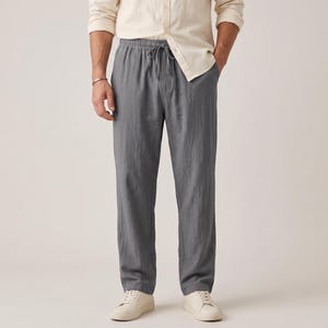Könnte beinhalten: Eine schiefergraue Hose mit elastischem Bund und passendem Kordelzug. Die Hose ist aus einem strukturierten Stoff gefertigt und wird mit einem hellbeigen Hemd mit Knopfleiste sowie weißen Turnschuhen kombiniert.