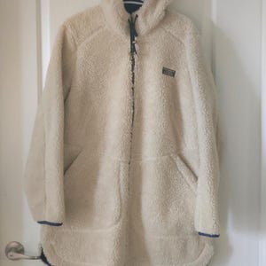 May include: A cream-coloured, long-sleeve fleece jacket with a textured, sherpa-like exterior, a hood, and a full-length front zip. The jacket features dark blue trim along the hem, cuffs, and zip, with a small rectangular logo on the chest.