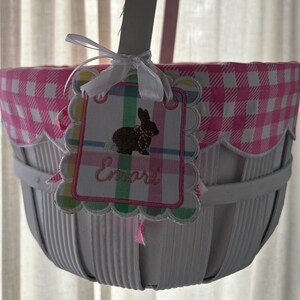 Op de afbeelding: Witte paasmand met een roze en witte ruitjesvoering. Een label met een konijnenillustratie en de naam "Emare" is bevestigd met een wit lint. De mand heeft een geschulpte rand en een handvat.