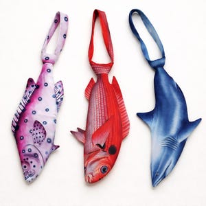 Könnte beinhalten: Drei ausgefallene Krawatten in Fischform. Eine ist rosa mit blauen Punkten, eine andere ist rot und die dritte ist blau und hat die Form eines Hais. Jede Krawatte hat oben eine Schlaufe zum Tragen.