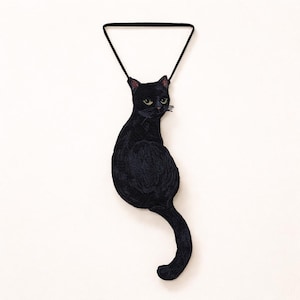 Peut inclure: Décoration murale en forme de chat noir, suspendue par un cordon noir triangulaire. Le chat est représenté assis, la queue recourbée. Les yeux du chat sont jaunes et il a un aspect texturé et détaillé.