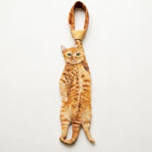 Könnte beinhalten: Eine ausgefallene Krawatte mit einem realistischen Bild einer stehenden, orangefarbenen Tabby-Katze. Die Krawatte hat einen hellbraunen Knoten und eine Schlaufe, und das Bild der Katze ist auf den Körper der Krawatte gedruckt. Die Katze hat grüne Augen und einen neugierigen Ausdruck.