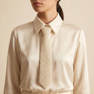 Può includere: Una cravatta realizzata interamente con piccole perle artificiali rotonde color crema disposte in un fitto e uniforme motivo a griglia. La cravatta è indossata sopra una camicia a maniche lunghe abbottonata in raso liscio e lucido color crema.