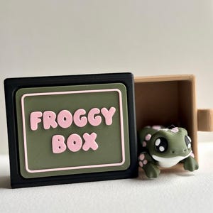 May include: A black and green box with the text "FROGGY BOX" in pink letters. A small green frog figurine with pink spots is inside a wooden box. The frog has large black eyes.