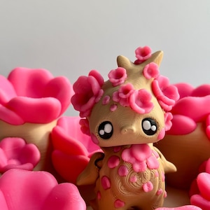 May include: A tan figurine with pink flower accents and large black eyes. The figurine is surrounded by pink flower-shaped objects. The design has a whimsical, fantasy theme.