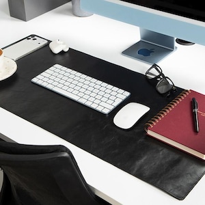 Può includere: Un'impostazione moderna della scrivania con un tappetino da scrivania in pelle nera, una tastiera e un mouse bianchi e un monitor del computer. Una tazza di caffè, un taccuino e occhiali sono anche sulla scrivania.
