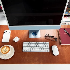 Puede incluir: Un tapete de escritorio de cuero marrón con una computadora, teclado, mouse, teléfono, taza de café y cuaderno. La computadora tiene un logotipo de Apple azul. El café tiene arte latte.