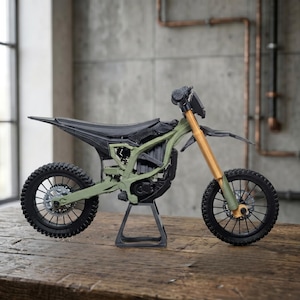 May include: A miniature dirt bike model featuring an olive green frame, black seat and fenders, and gold-colored front suspension forks. The motorcycle is supported by a black stand and has black spoked wheels with detailed brake discs.