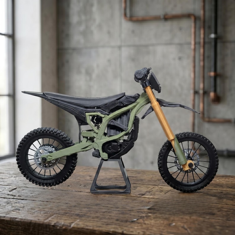 May include: A miniature dirt bike model featuring an olive green frame, black seat and fenders, and gold-colored front suspension forks. The motorcycle is supported by a black stand and has black spoked wheels with detailed brake discs.