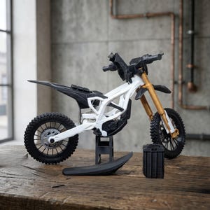 May include: A white and black dirt bike model with gold-colored front forks, black knobby tires, and a black seat. The motorcycle is positioned on a small black stand, with a separate black fender and a rectangular black battery pack placed nearby.