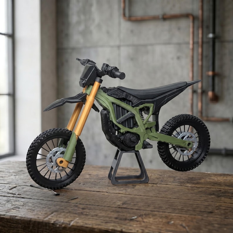 May include: A miniature dirt bike model featuring an olive green frame, black seat and fenders, and gold-colored front suspension forks. The motorcycle is supported by a black stand and includes black knobby tires with silver disc brakes.