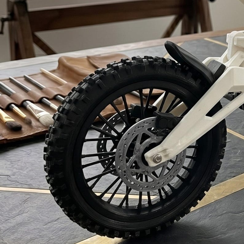 May include: A black motorcycle wheel with a knobby tire and a silver disc brake rotor attached to a white frame. The wheel features thin black spokes and a black mudguard positioned above the tire, showcasing detailed mechanical components.