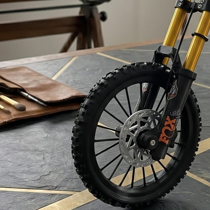 May include: A miniature bicycle front wheel assembly featuring a black knobby tire, black spokes, and a silver disc brake rotor. The suspension forks are gold-colored with black lower sections, displaying the orange Fox brand logo and text.