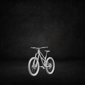 May include: A white mountain bike with chunky tyres and a suspension fork. The bike features a curved frame, a seat, and handlebars. The wheels have a detailed tread pattern. The background is a dark, textured surface.