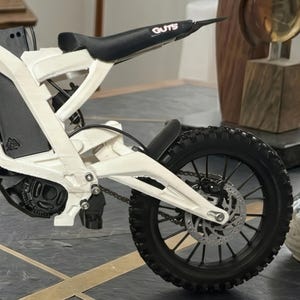 May include: A white electric dirt bike model featuring a black seat with the text GUTS printed in white. The bike has a black rear tire with a disc brake, a chain drive system, and a black motor housing. The frame is white with visible suspension.