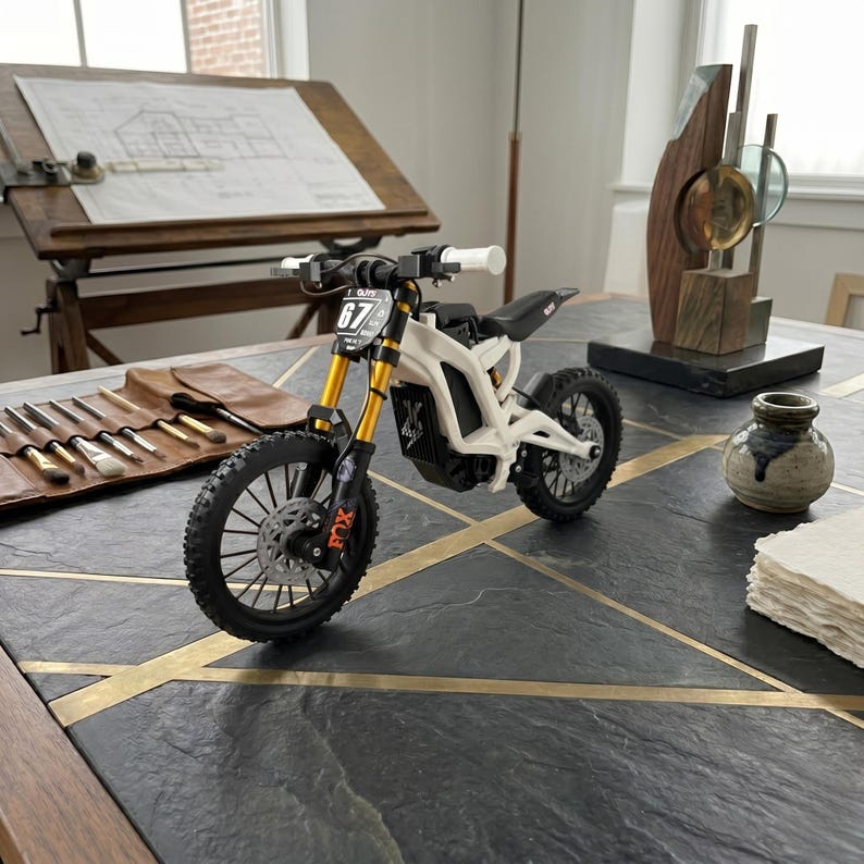 May include: A miniature electric dirt bike model featuring a white frame, gold-colored front suspension forks, black knobby tires, and a black seat. The front number plate displays the number 67, and the front fork includes orange Fox branding.
