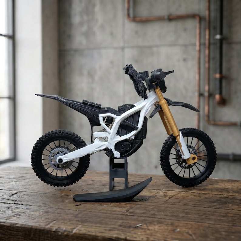 May include: A miniature dirt bike model featuring a white frame, black seat and tires, and gold-colored front suspension forks. The motorcycle is supported by a small black stand and includes detailed components like a rear sprocket and disc brakes.