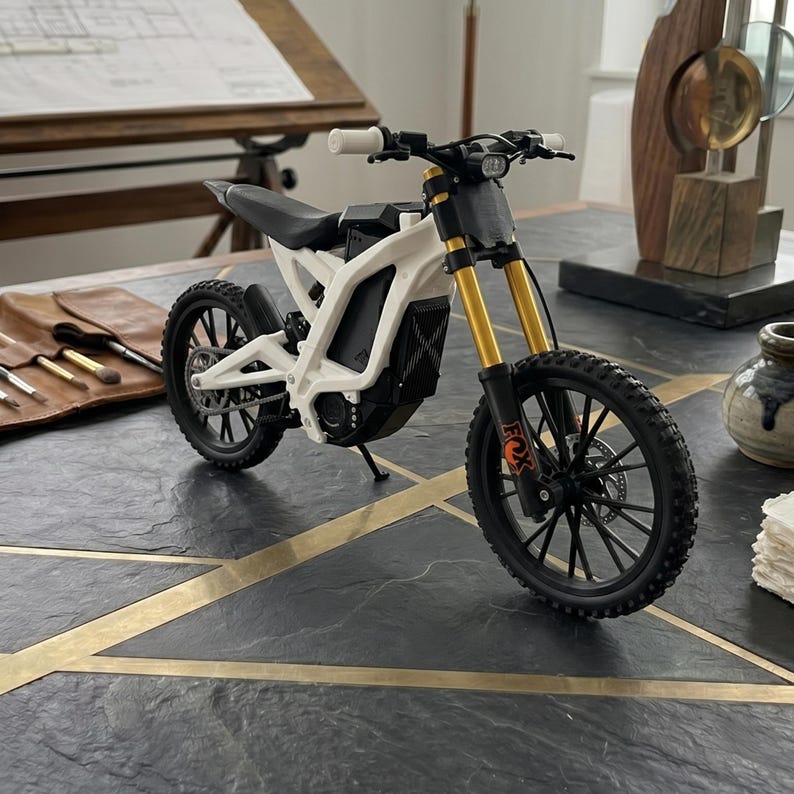 May include: A white and black electric dirt bike model featuring gold-colored front suspension forks, black spoked wheels with knobby tires, a black seat, and a small headlight. The frame is white with a black central battery housing and motor.