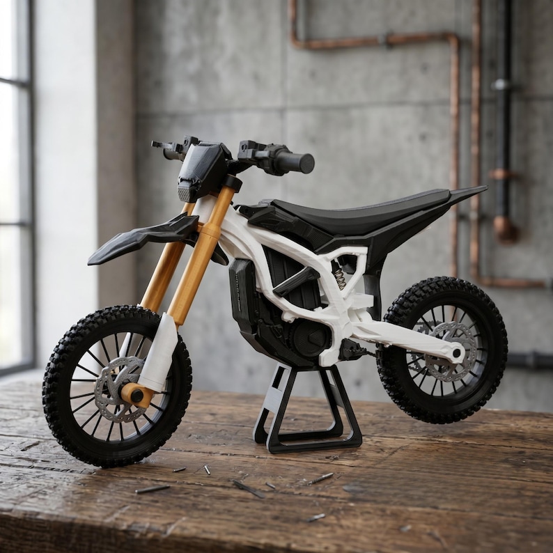 May include: A miniature dirt bike model featuring a white frame, black seat, and black handlebars. The front suspension forks are gold, and the bike is supported by a black stand. It has black knobby tires with silver disc brakes.