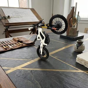 May include: A miniature white and black electric dirt bike model performing a wheelie. The bike features gold-colored front suspension forks, black spoked wheels with knobby tires, and a small number plate on the handlebars with the number 67 visible.