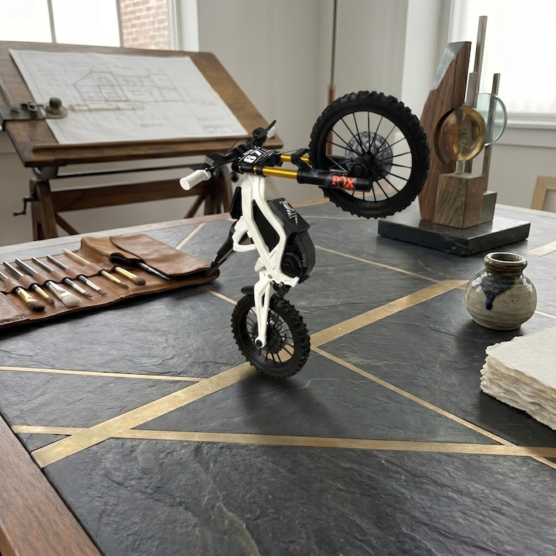 May include: A miniature white and black electric dirt bike model performing a wheelie. The bike features gold-colored front suspension forks, black spoked wheels with knobby tires, and a small number plate on the handlebars with the number 67 visible.