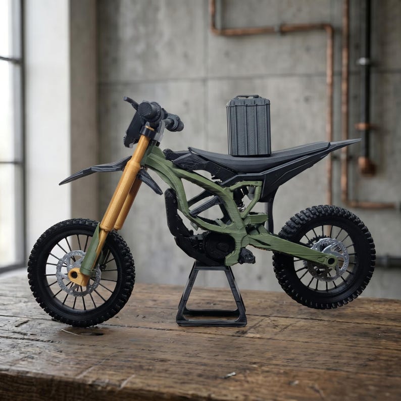 May include: A miniature olive green dirt bike model featuring gold-colored front forks, black spoked wheels with disc brakes, a black seat, and a vertical grey battery pack mounted on the frame, all supported by a black metal display stand.