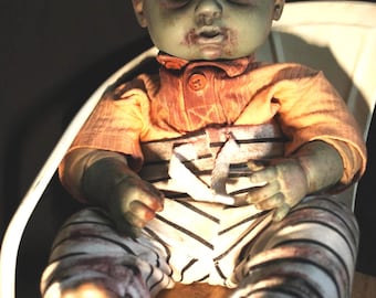 May include: A doll named Toby with pale green skin, dark sunken eyes, and red-stained lips. The doll wears an orange collared shirt and white pants with thin black horizontal stripes. The doll has a weathered appearance with visible dark smudges.