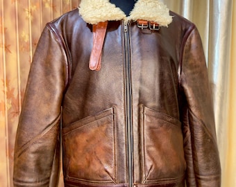May include: Brown leather bomber jacket with a cream-colored shearling collar and cuffs. The jacket features a zip-up front, two front pockets, and a buckle detail at the collar. The jacket is displayed on a mannequin.