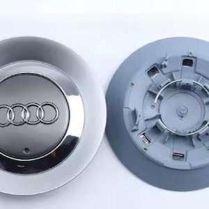 May include: Two silver and grey Audi car wheel centre caps. One cap features the Audi logo of four interlocking rings. The other cap is grey and shows the back side with a metal ring.