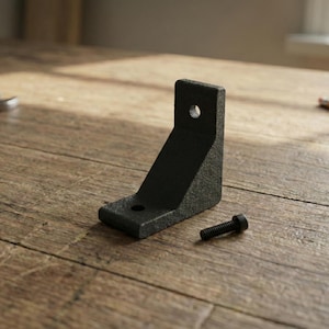 May include: A black metal corner bracket with two pre-drilled holes and a matching black screw. The bracket is angled at 90 degrees and sits on a wooden surface. The image is well-lit, highlighting the texture of the bracket.