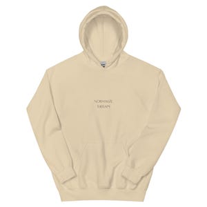 May include: A beige hoodie with a front pocket and a drawstring hood. The text "NORMALIZE THERAPY" is printed in a small, centered font on the front. The sweatshirt is made of a soft material. A casual garment.