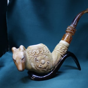 May include: A carved tobacco pipe with a camel head design. The pipe is beige with brown accents and rests on a black stand. The bowl features intricate carvings, and the stem is curved.