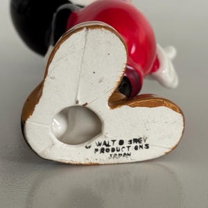 May include: A ceramic Mickey Mouse figurine, viewed from the bottom up. The figure has a red torso, black head and ears, and brown feet. The text "WALT DISNEY PRODUCTIONS JAPAN" is printed on the bottom of the foot.