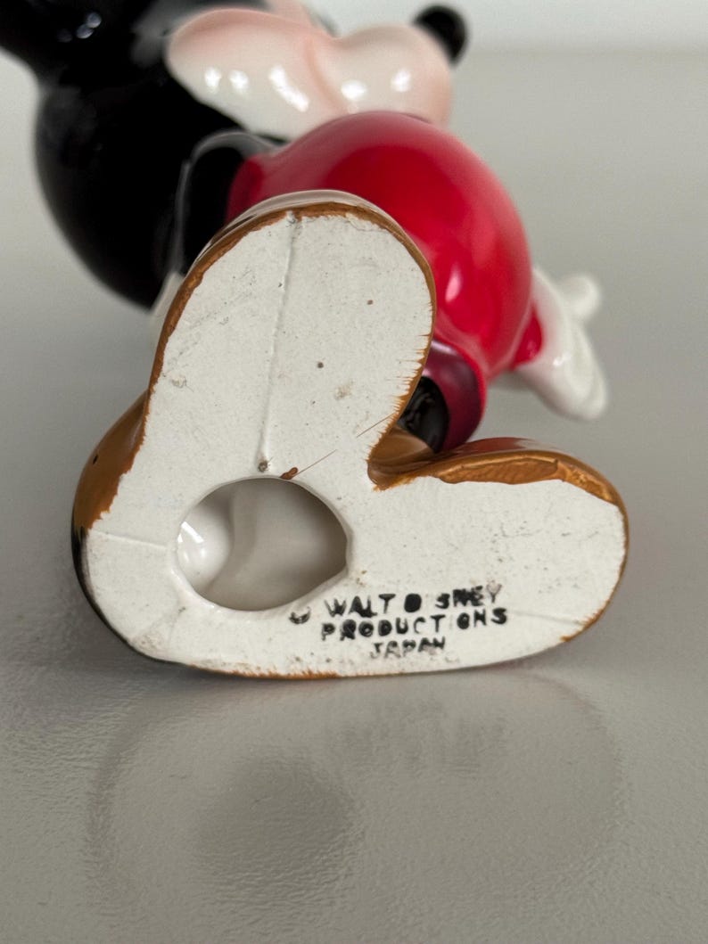 May include: A ceramic Mickey Mouse figurine, viewed from the bottom up. The figure has a red torso, black head and ears, and brown feet. The text "WALT DISNEY PRODUCTIONS JAPAN" is printed on the bottom of the foot.