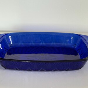 May include: A rectangular, cobalt blue glass baking dish with rounded handles. The dish features a leaf pattern around the base and a textured design on the interior. The dish is empty and sits on a white surface.