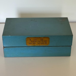 May include: A rectangular, teal-colored metal storage box with a hinged lid. A gold-toned rectangular label is centered on the front, featuring the black text "The American Home MENU MAKER" in a classic serif font. The box shows minor surface wear.