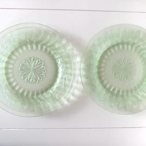 May include: Two round, light green glass plates featuring a central embossed floral medallion pattern surrounded by a ring of vertical ridges and an outer border decorated with intricate, swirling leaf and flower motifs in relief.