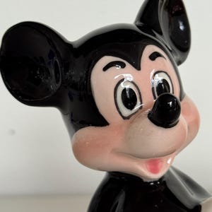 May include: A ceramic Mickey Mouse figurine. The figure has a black head and ears, a pink face, and a black nose. The figurine is wearing a black body with a red bottom. The figurine is a classic Disney character.