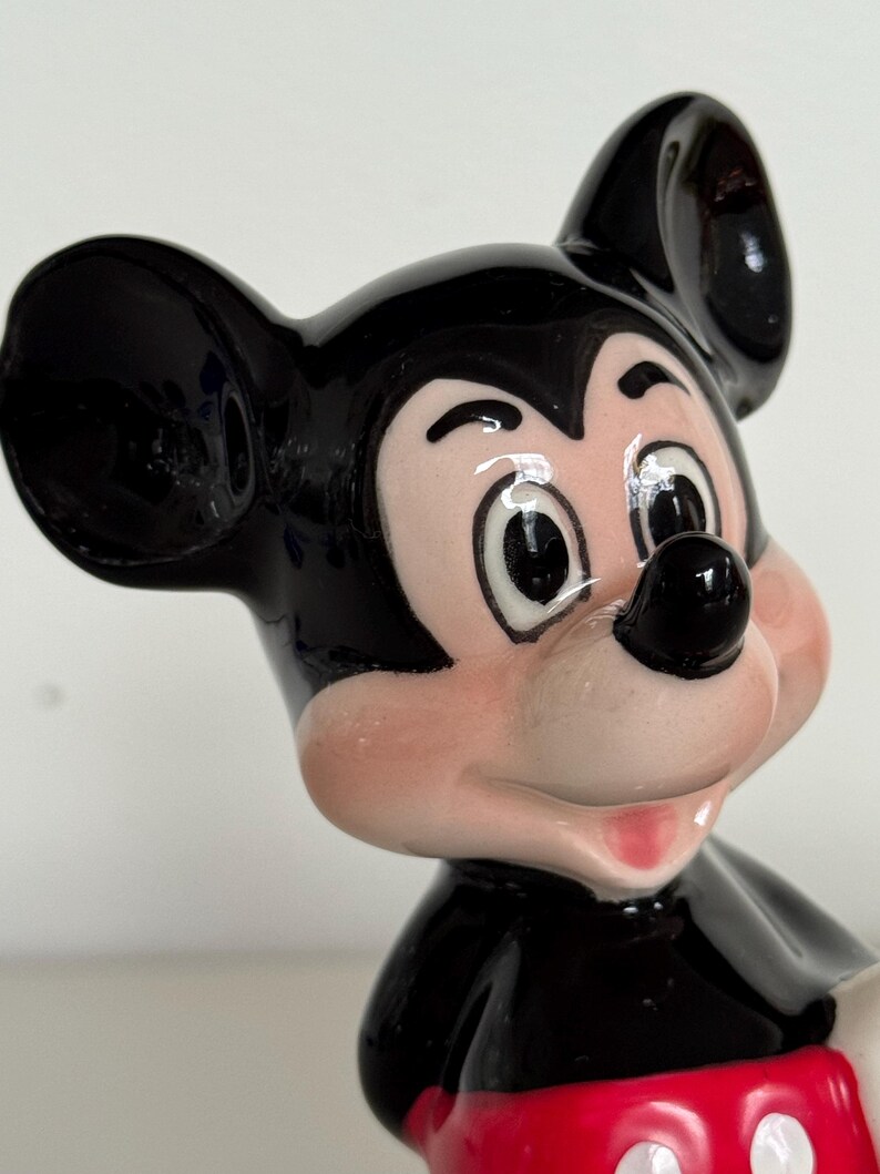 May include: A ceramic Mickey Mouse figurine. The figure has a black head and ears, a pink face, and a black nose. The figurine is wearing a black body with a red bottom. The figurine is a classic Disney character.