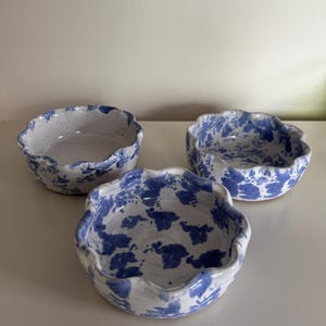 May include: Three ceramic bowls with a white base and blue splatter pattern. The bowls have a wavy, scalloped rim. The bowls are of varying sizes and are arranged on a white surface.