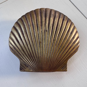 May include: A brass scallop shell dish featuring a metallic gold finish with a textured, ribbed surface. The object is shaped like a natural seashell with a fan-like design, radiating from the base to the scalloped edge, showing a weathered patina.