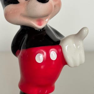 May include: A ceramic Mickey Mouse figurine. The figure has a black body, red shorts with white buttons, and white gloves. The figurine is smiling and giving a thumbs-up. A collectible item.