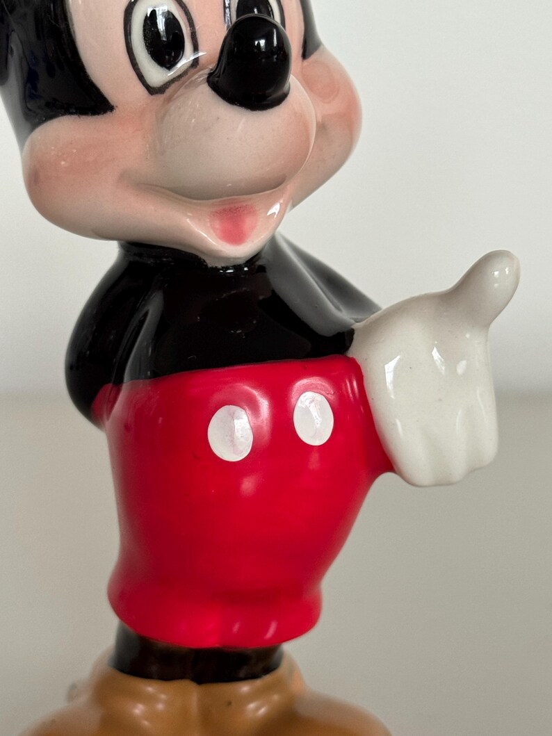 May include: A ceramic Mickey Mouse figurine. The figure has a black body, red shorts with white buttons, and white gloves. The figurine is smiling and giving a thumbs-up. A collectible item.