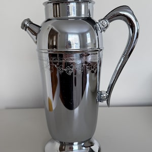 May include: A polished silver cocktail shaker with a lid, spout, and handle. The shaker has a decorative etched design around the middle. The handle is curved and attached to the body of the shaker. The shaker sits on a round base.