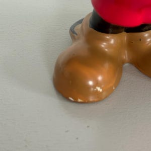 May include: Close-up of a cartoon character's brown shoe with a red garment above. The shoe has a rounded toe and a glossy finish. The shoe is a light brown colour with some white paint scuffs.