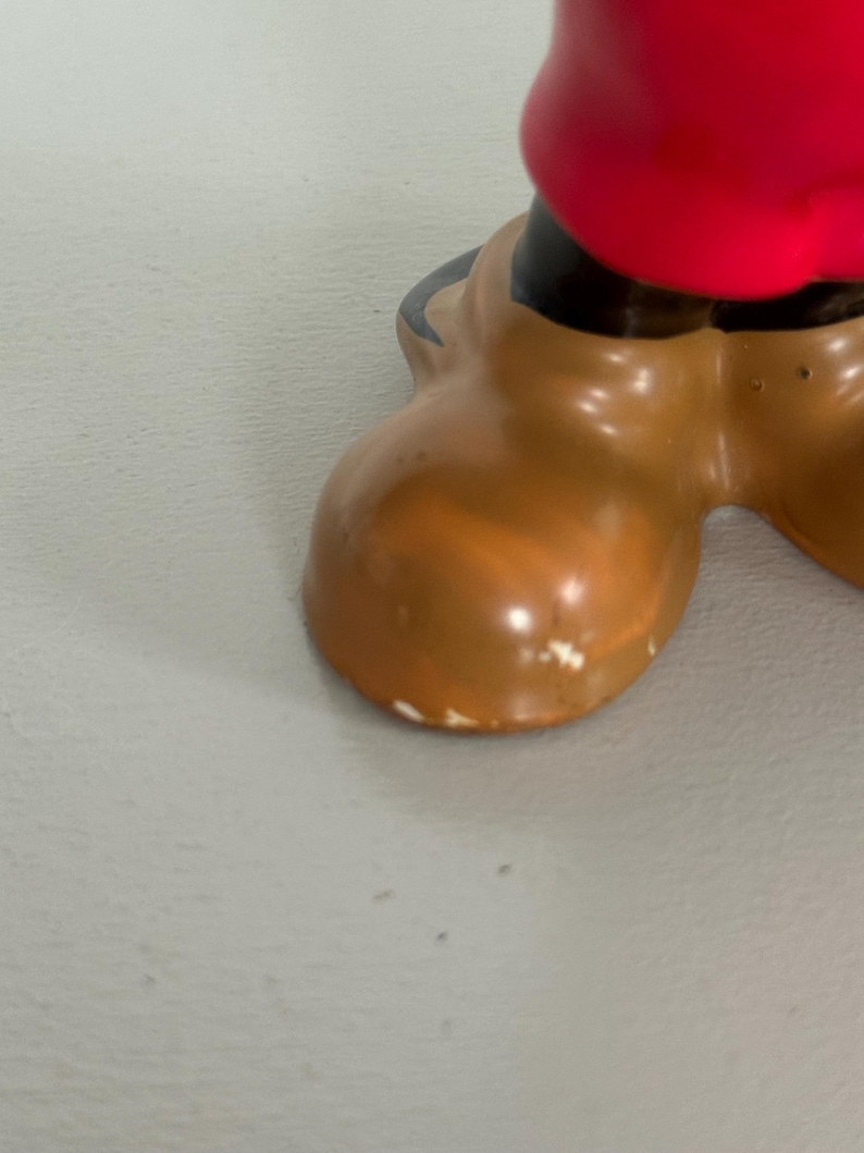 May include: Close-up of a cartoon character's brown shoe with a red garment above. The shoe has a rounded toe and a glossy finish. The shoe is a light brown colour with some white paint scuffs.