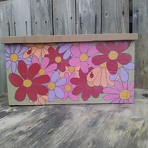 May include: A rectangular wooden box with a light brown lid and a floral design. The box is painted with large flowers in shades of red, pink, purple, and orange, with yellow centers. A small insect is painted on the box.