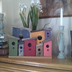 May include: A multi-unit wooden birdhouse structure featuring seven individual compartments painted in various colors including blue, green, red, orange, and purple, all mounted on a single rectangular wooden base with circular entry holes.