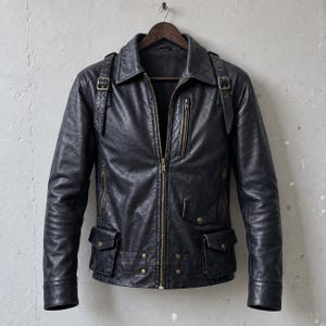 Könnte beinhalten: Eine schwarze Lederjacke mit zentralem Reißverschluss, einer vertikalen Reißverschlusstasche auf der Brust und zwei unteren Pattentaschen mit Druckknöpfen. Die Jacke verfügt über Schulterriegel mit Metallschnallen und einen Gürtel mit Nieten.